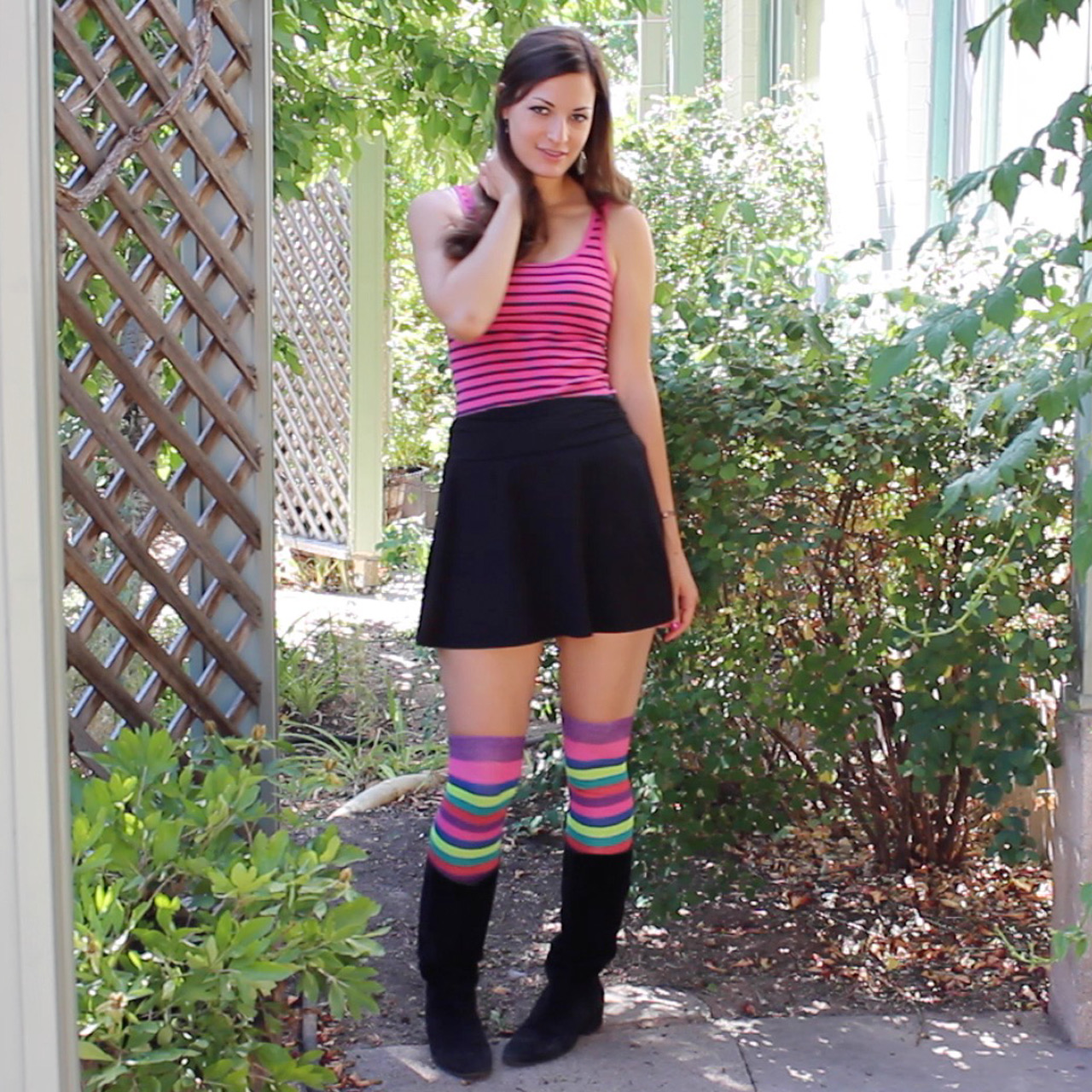 A woman standing in a garden pathway wearing a pink and black striped tank top, black skirt, striped long socks in shades of purple, blue, and green, and black mid-calf boots.