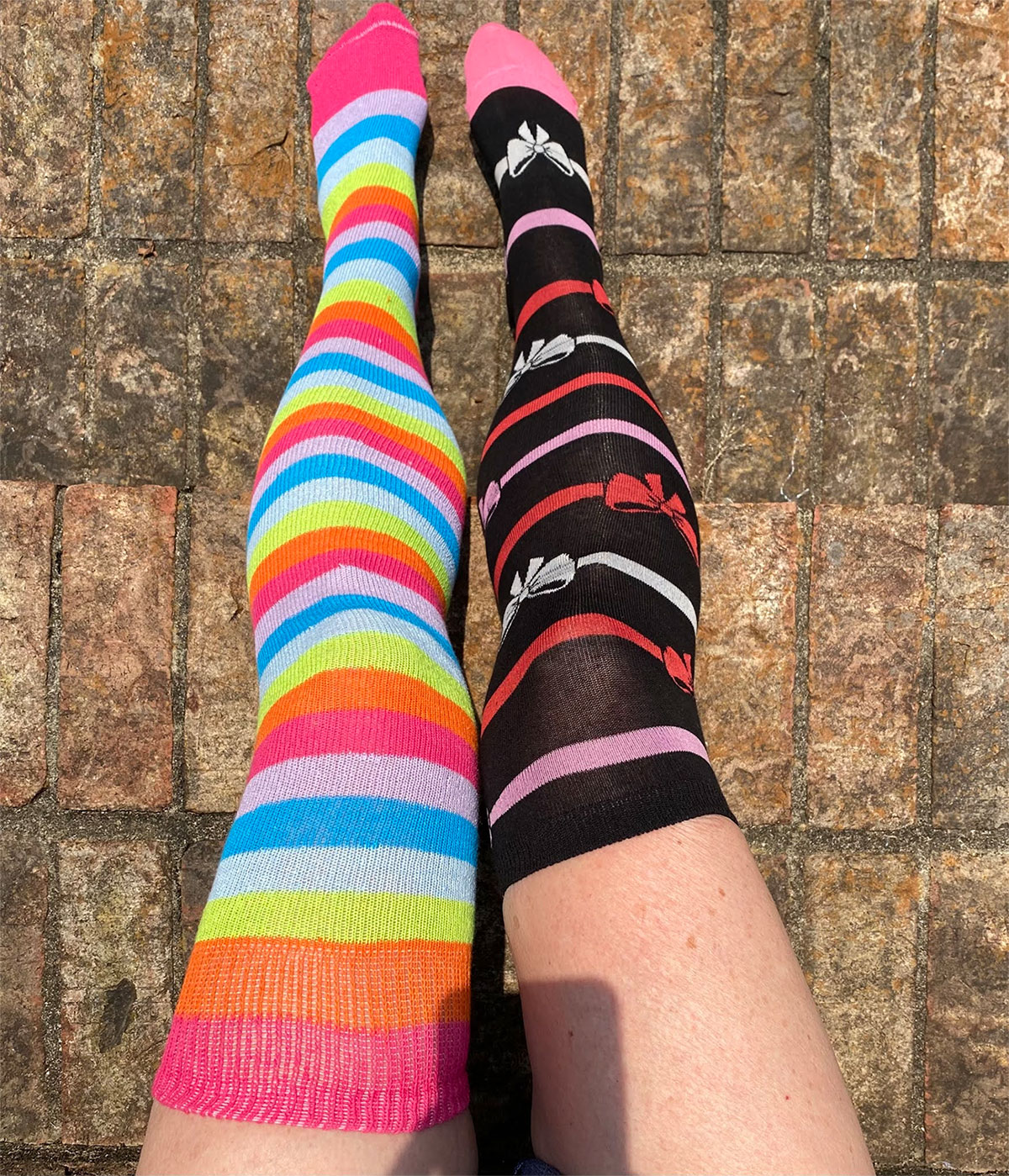 2 different pairs of socks, one long rainbow striped thigh high and the other a knee high socks with bow and ribbon creating a unique look