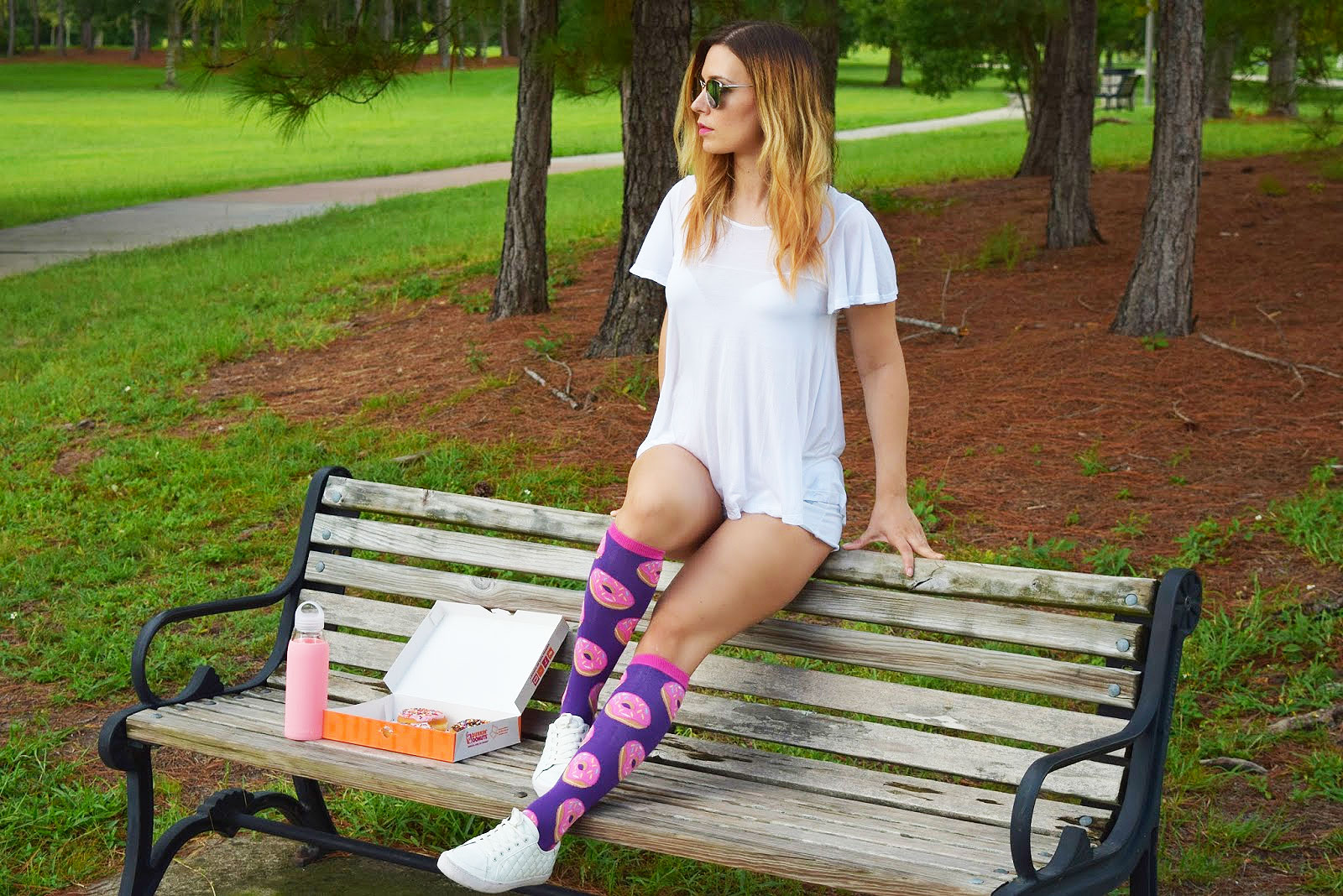 A woman sitting on a park bench in purple donut-themed socks, next to a box of donuts and a water bottle.