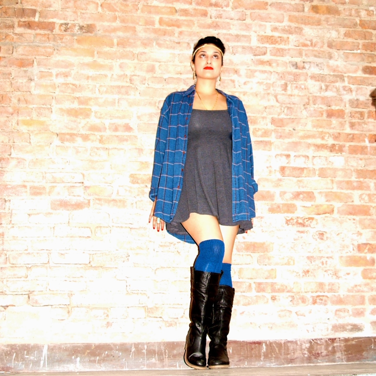 A girl in a casual pose against a brick wall backdrop, wearing a dark gray dress, blue thigh high socks, black leather boots, and a blue checkered shirt with rolled-up sleeves.