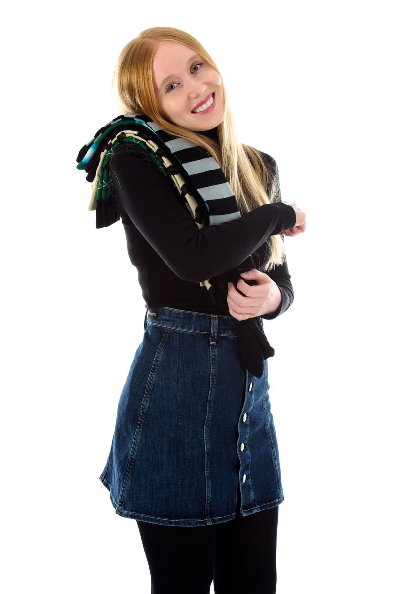 A smiling woman with blonde hair, wearing a black top, denim skirt, and black leggings, casually draped with various colorful striped socks over her shoulder, against a white background.