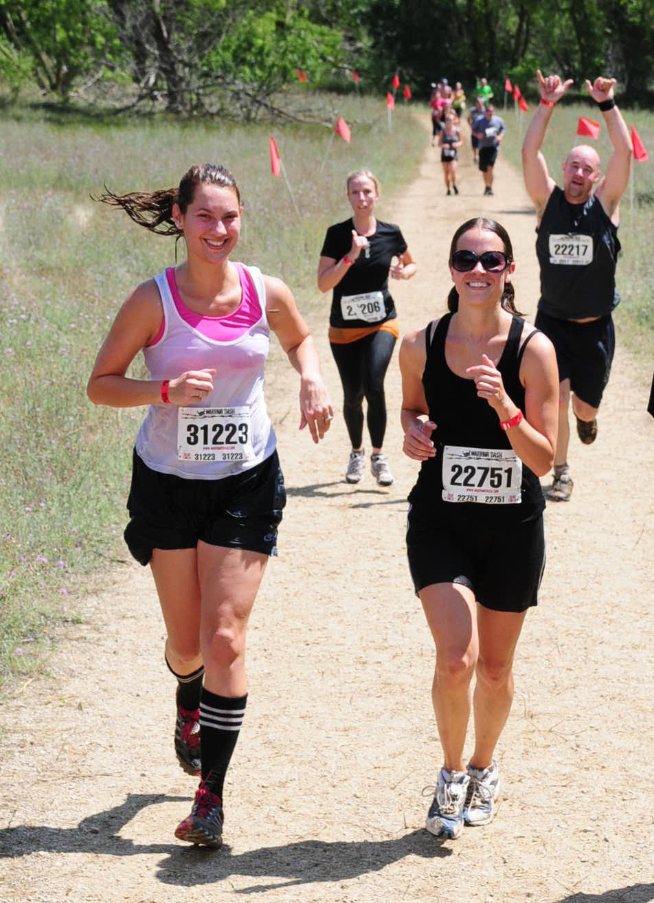 A group of runners participating in a mud run, smiling and showing enthusiasm as they compete. One runner in the foreground is wearing a white and pink tank top, black shorts, and knee-high athletic socks with distinctive black and white stripes, paired with running shoes.