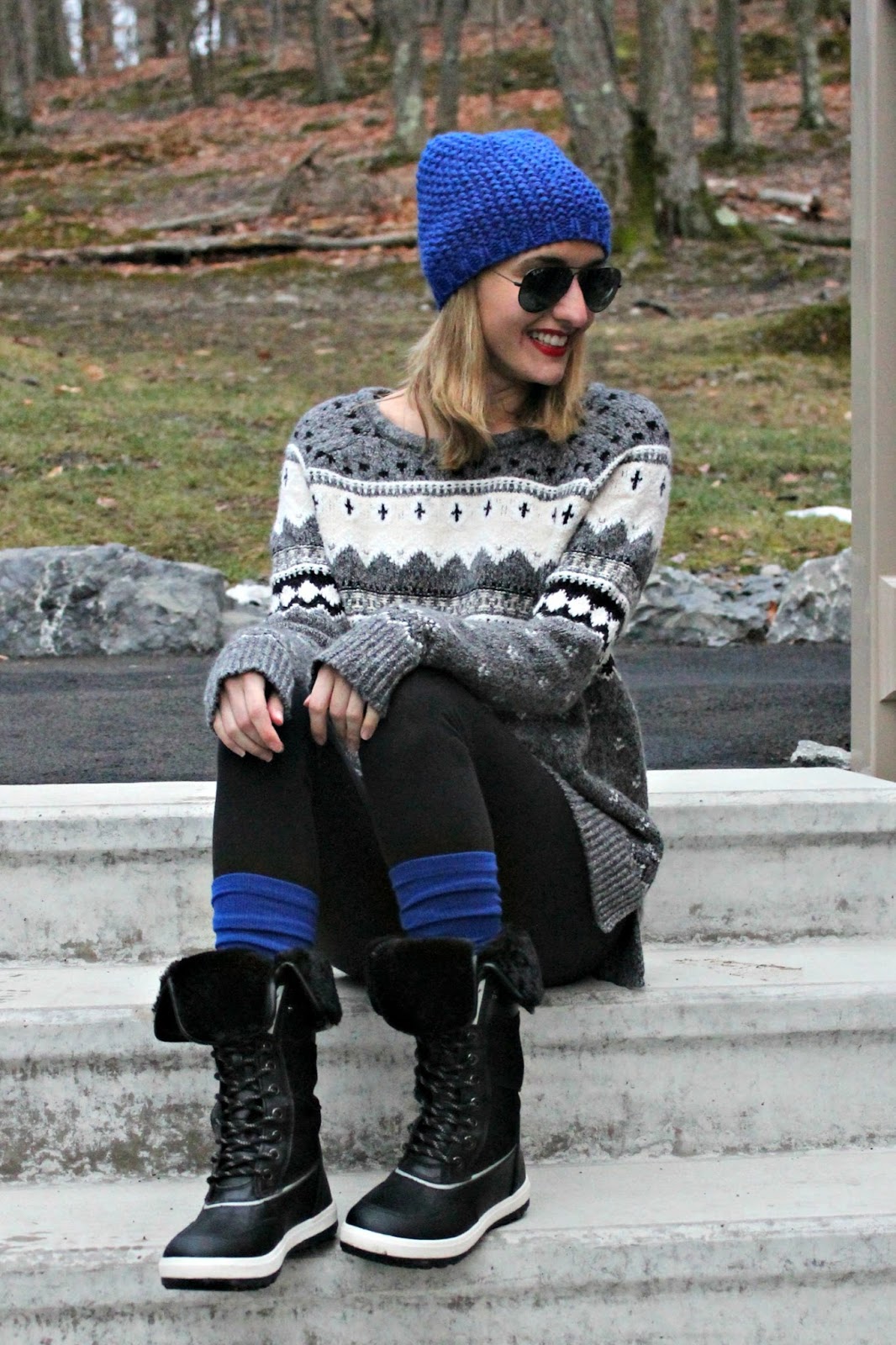 girl wearing black leggings, black winter boots, thick blue knee socks, a winter-themed sweater, a blue knit beanie, and black sunglasses