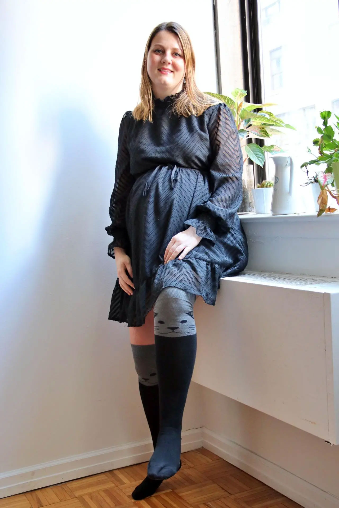 A woman standing by a window, wearing a black textured dress and gray knee-high socks with a cat face design.