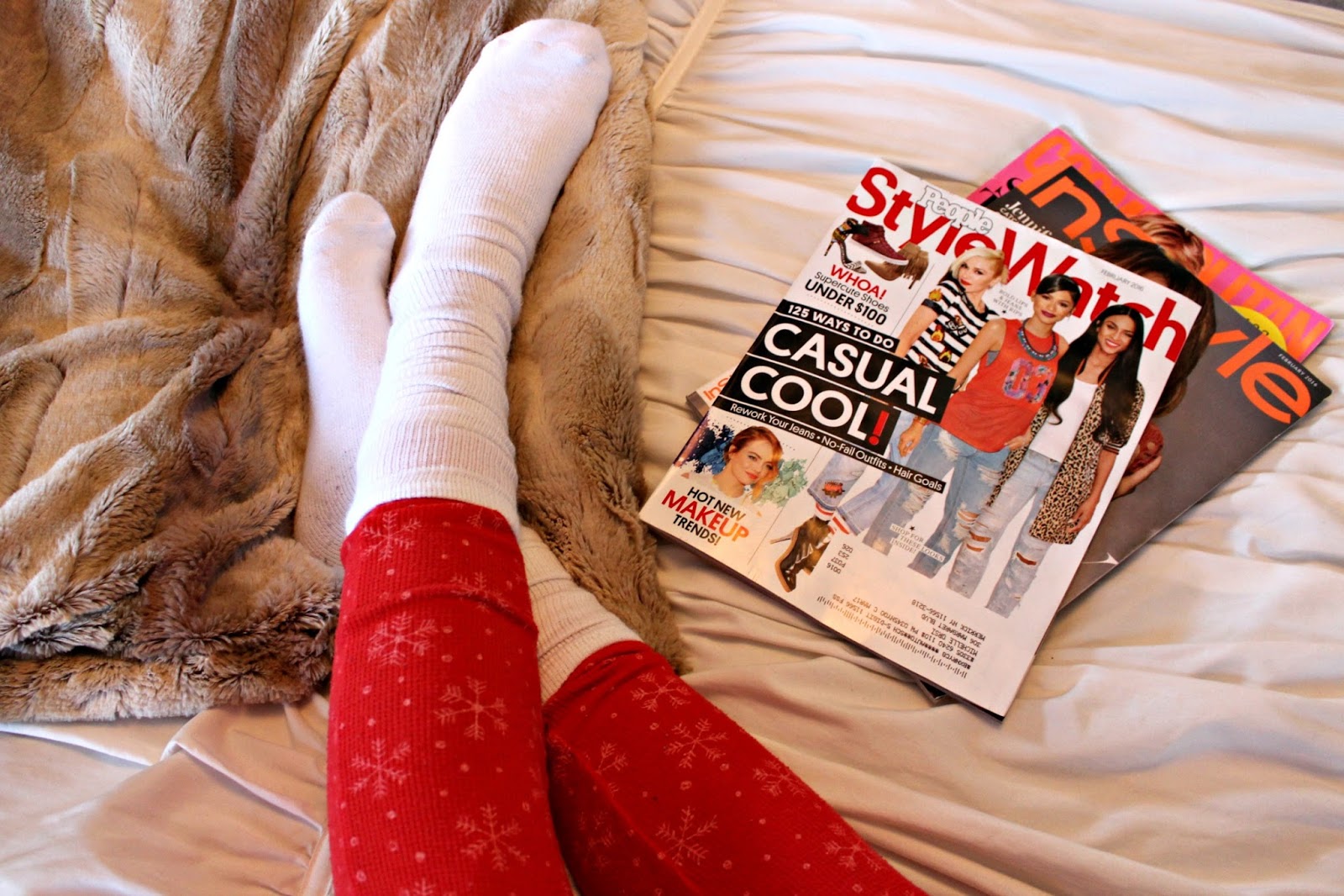A girl lying on a bed, reading magazines. She's wearing white socks and red pajamas with a snowflake pattern, resting on top of the sheets.