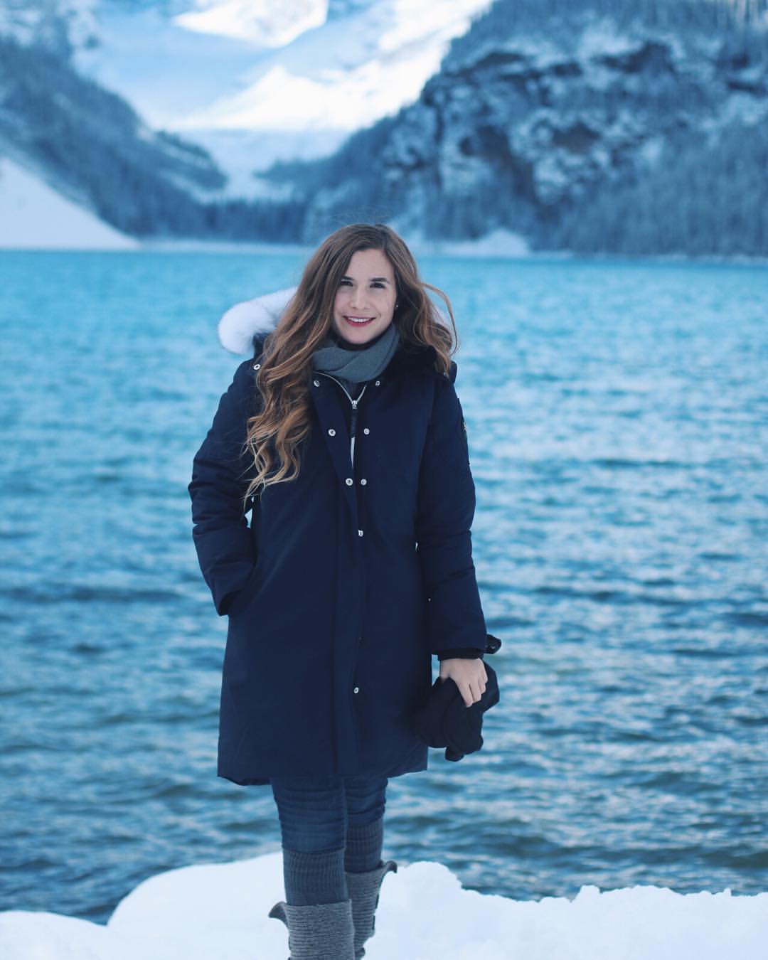 A woman in a navy coat and grey thigh-high socks stands before a snowy mountain landscape and a lake.