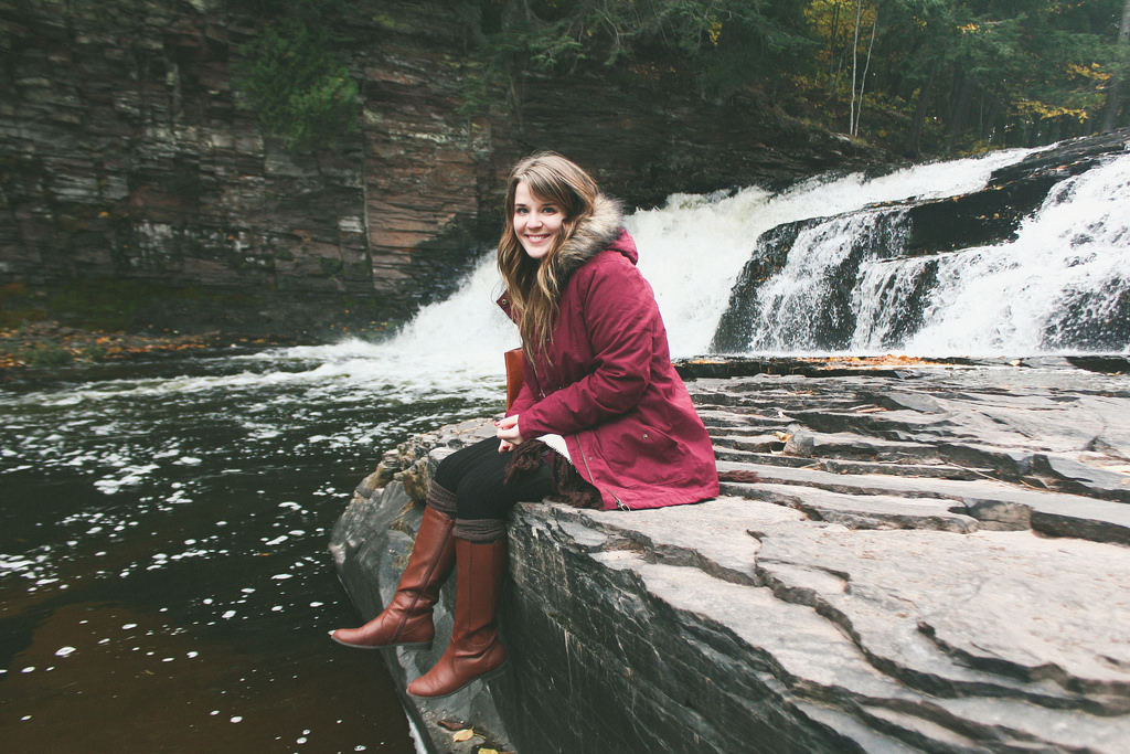A young woman in a maroon coat and brown boots sitting on layered rocks by a waterfall, with trees displaying late fall colors in the background.