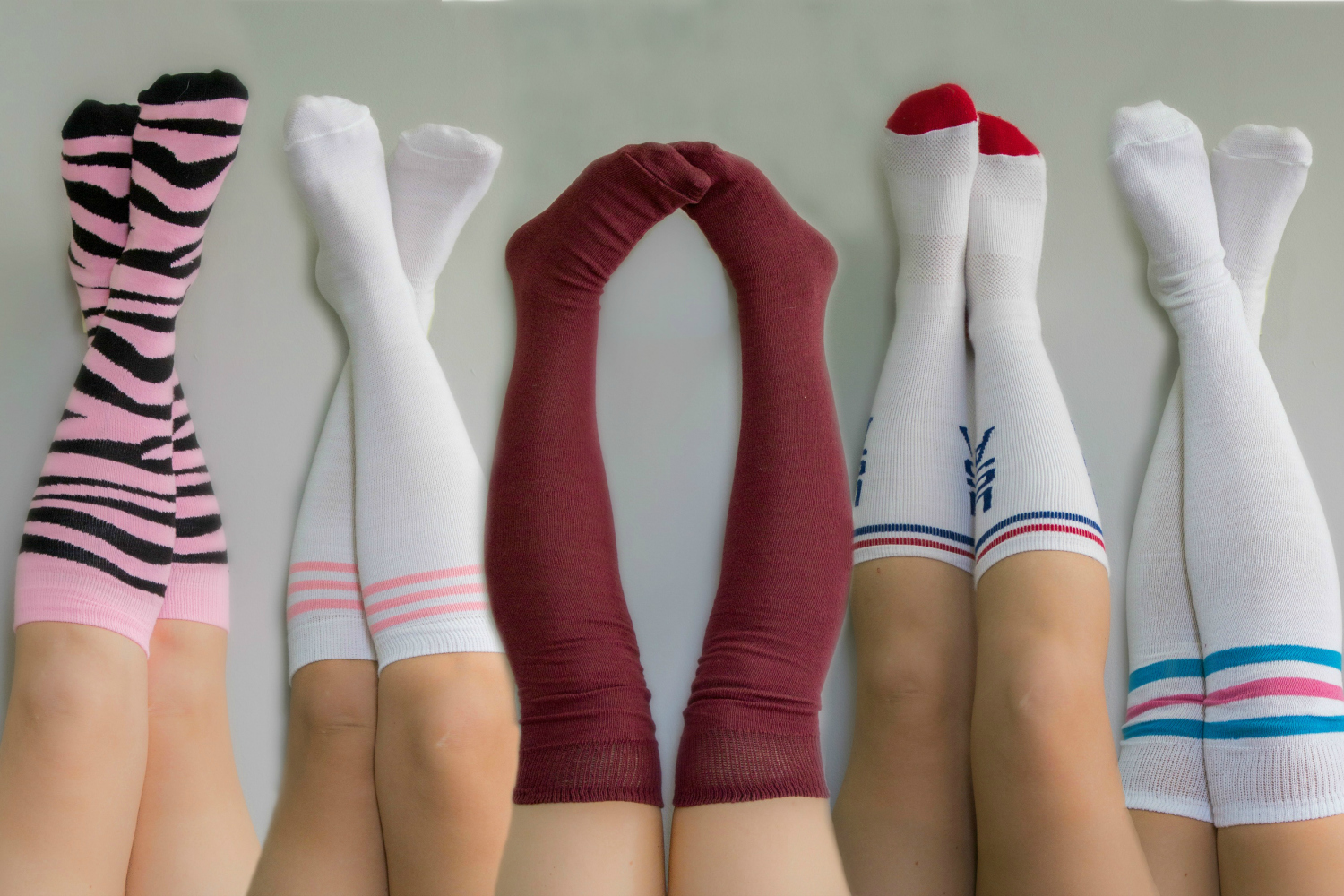 Five pairs of legs raised against a grey background, each donning unique knee-high socks, including zebra stripes, solid colors, and striped patterns