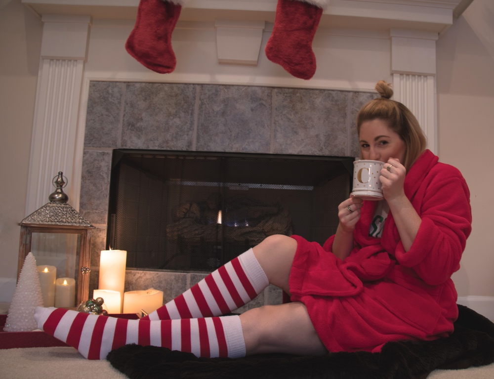 girl wearing red and white striped socks, a red robe, a white college-themed t-shirt, and shorts, drinking out of a coffee cup while sitting in front of a fireplace