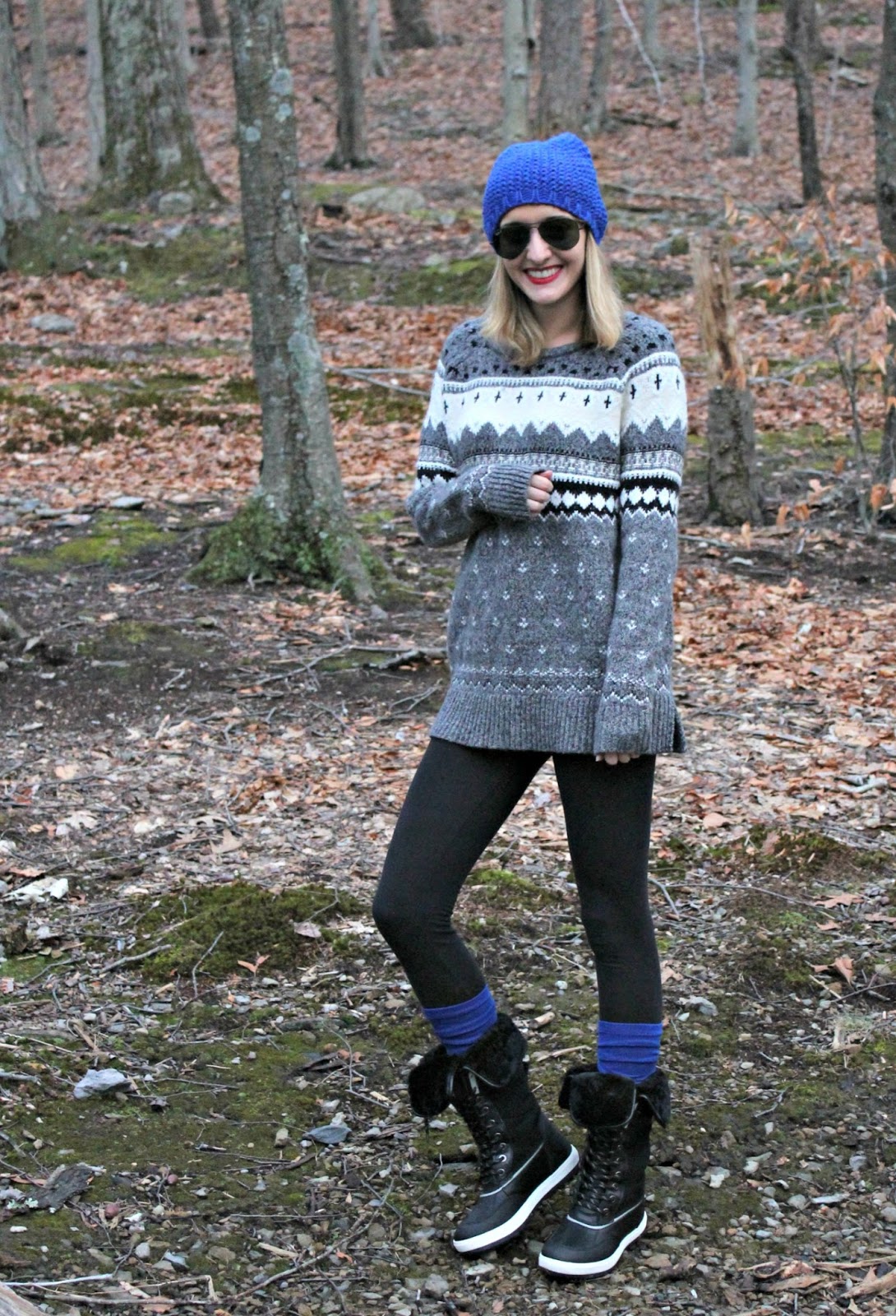 A smiling woman in a patterned sweater, black leggings, and winter boots with blue socks, wearing a blue beanie and sunglasses in a forest setting.