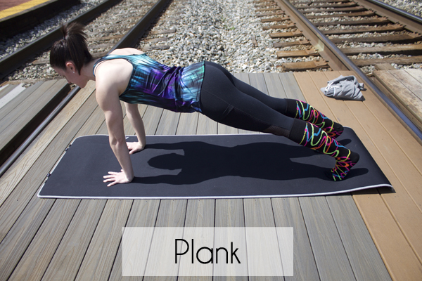 A girl performing a plank pose on a yoga mat placed on a wooden deck near railroad tracks.