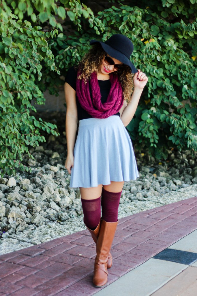 A young woman wearing a gray skater skirt, a black T-shirt, maroon knee-high socks, brown leather boots, a maroon scarf, and a black hat, posing playfully outdoors